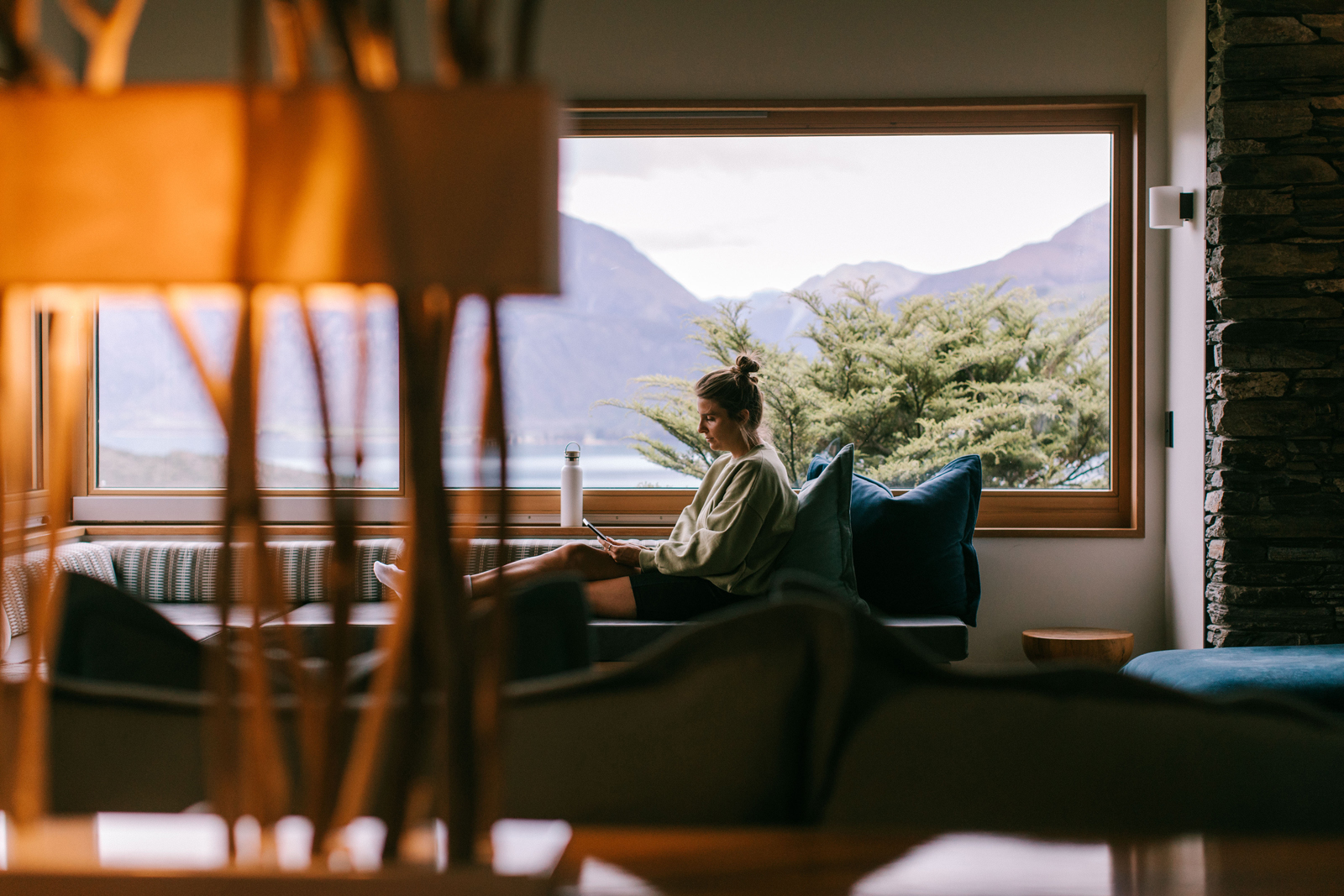 A person relaxes in an outdoor infinity pool with a breathtaking view of a serene mountain lake and snow-capped mountain ranges in the background.