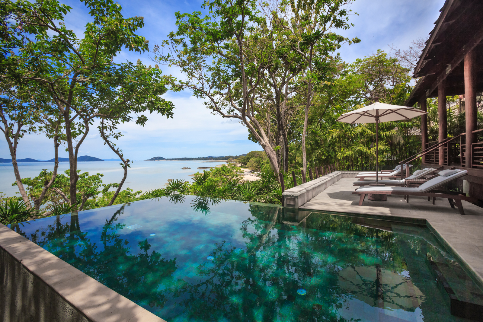 Kamalaya Koh Samui_Beach Front Pool Villa An infinity pool overlooking a serene tropical coastline, surrounded by lush trees and greenery with a stunning ocean and distant islands in the background. The pool area features comfortable loungers and an elegant open-air design.