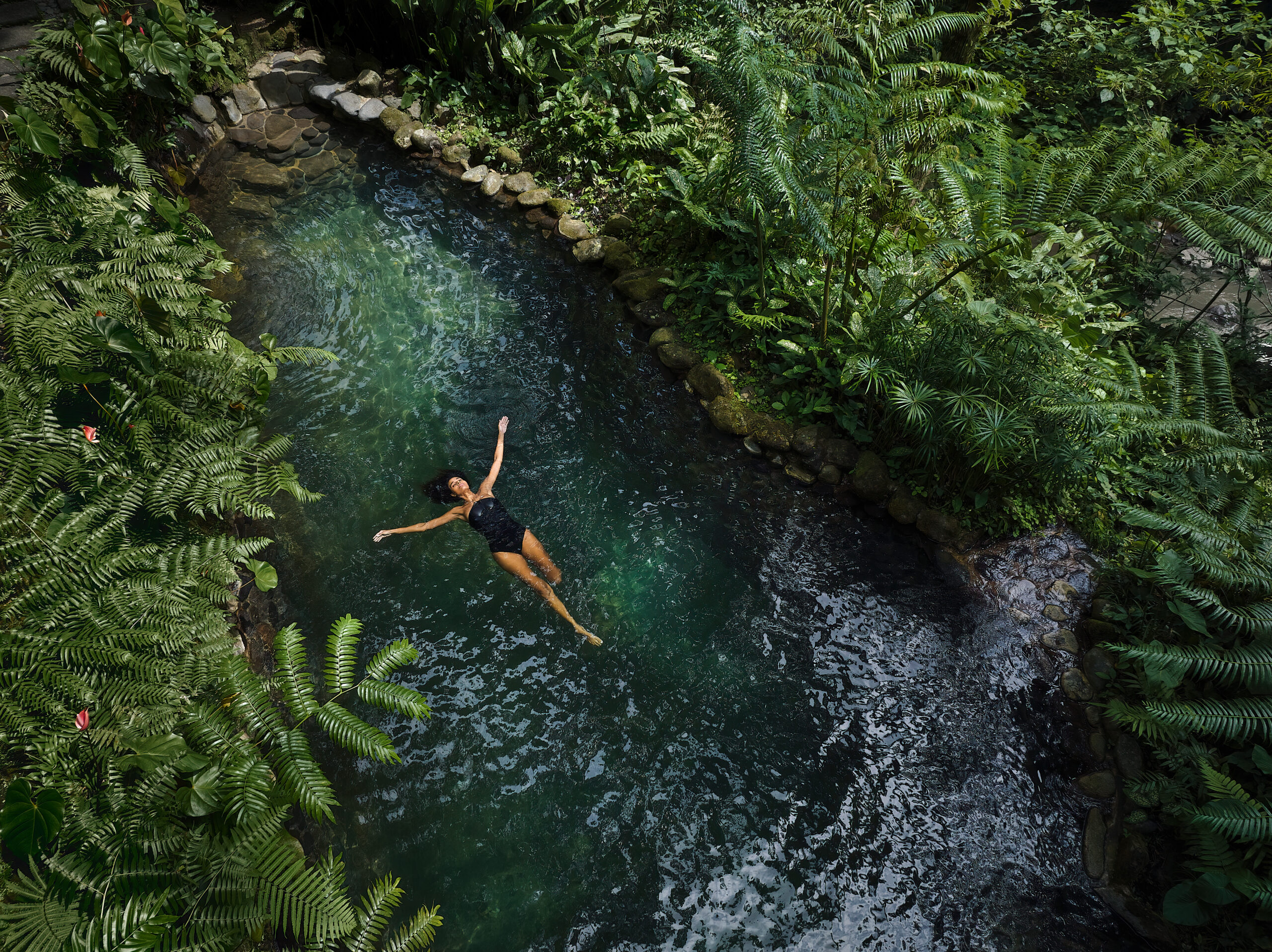 A person floating peacefully in a lush green jungle stream surrounded by ferns and tropical vegetation.