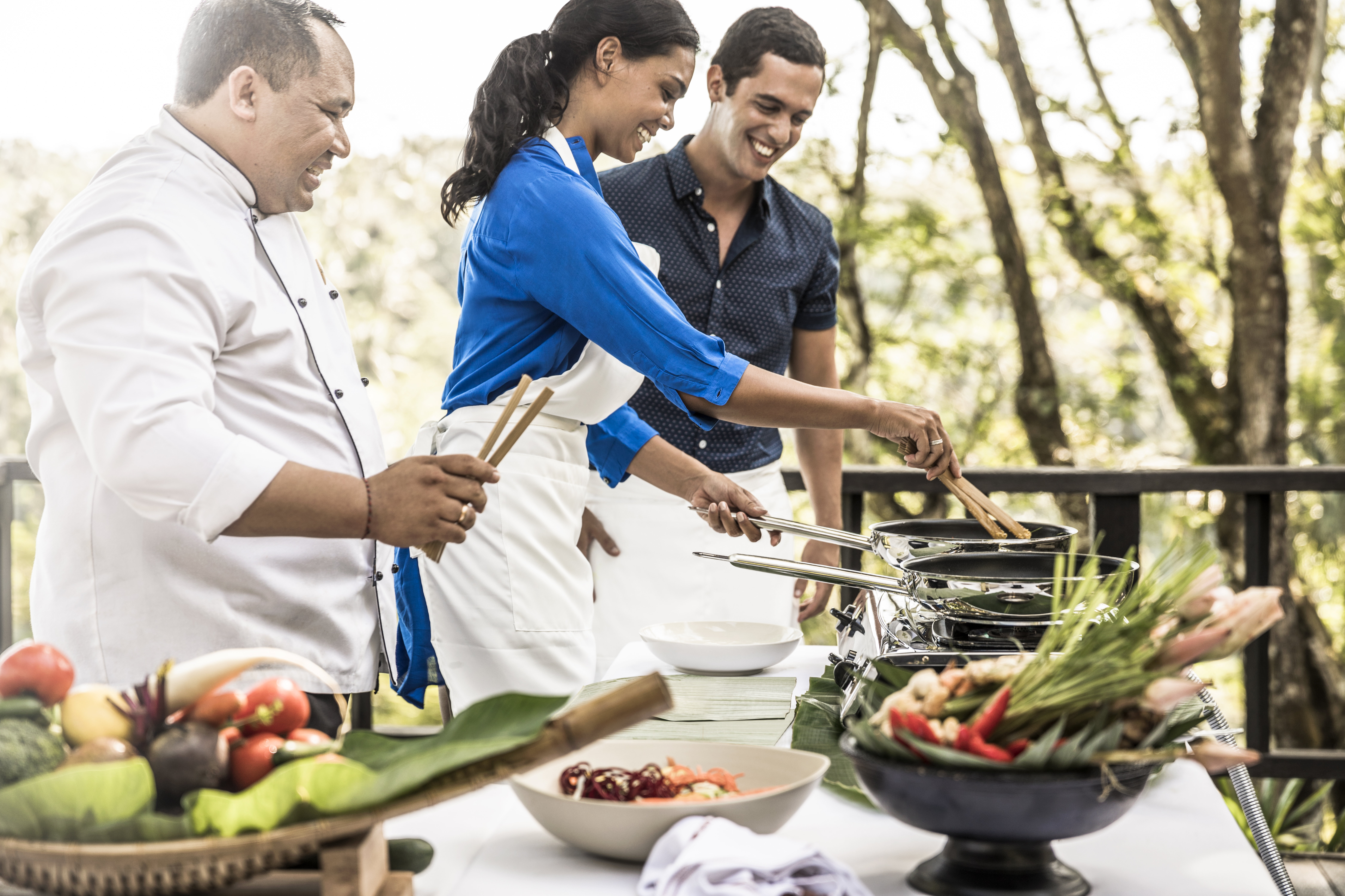 A chef and two people preparing food together on an outdoor grill surrounded by fresh vegetables and ingredients.