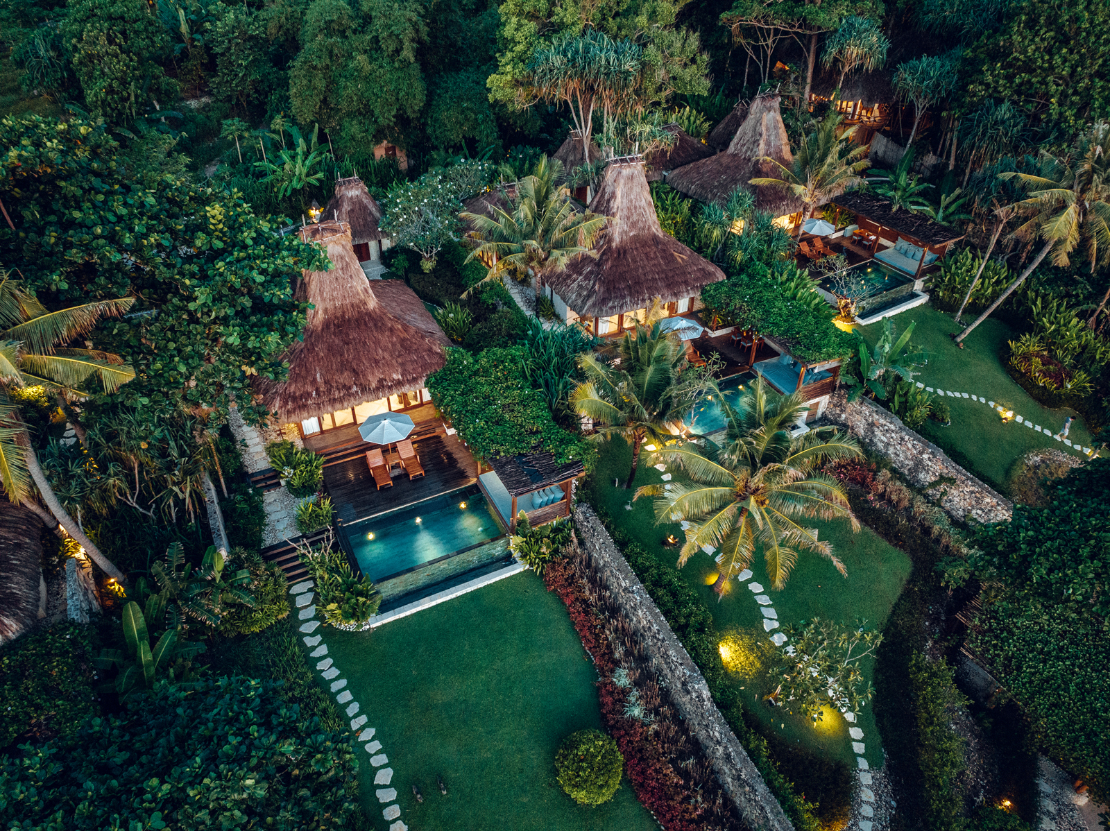 An aerial view of a lush tropical resort featuring traditional thatched-roof villas nestled among palm trees and landscaped gardens. The scene includes private swimming pools and illuminated pathways connecting the various accommodations.