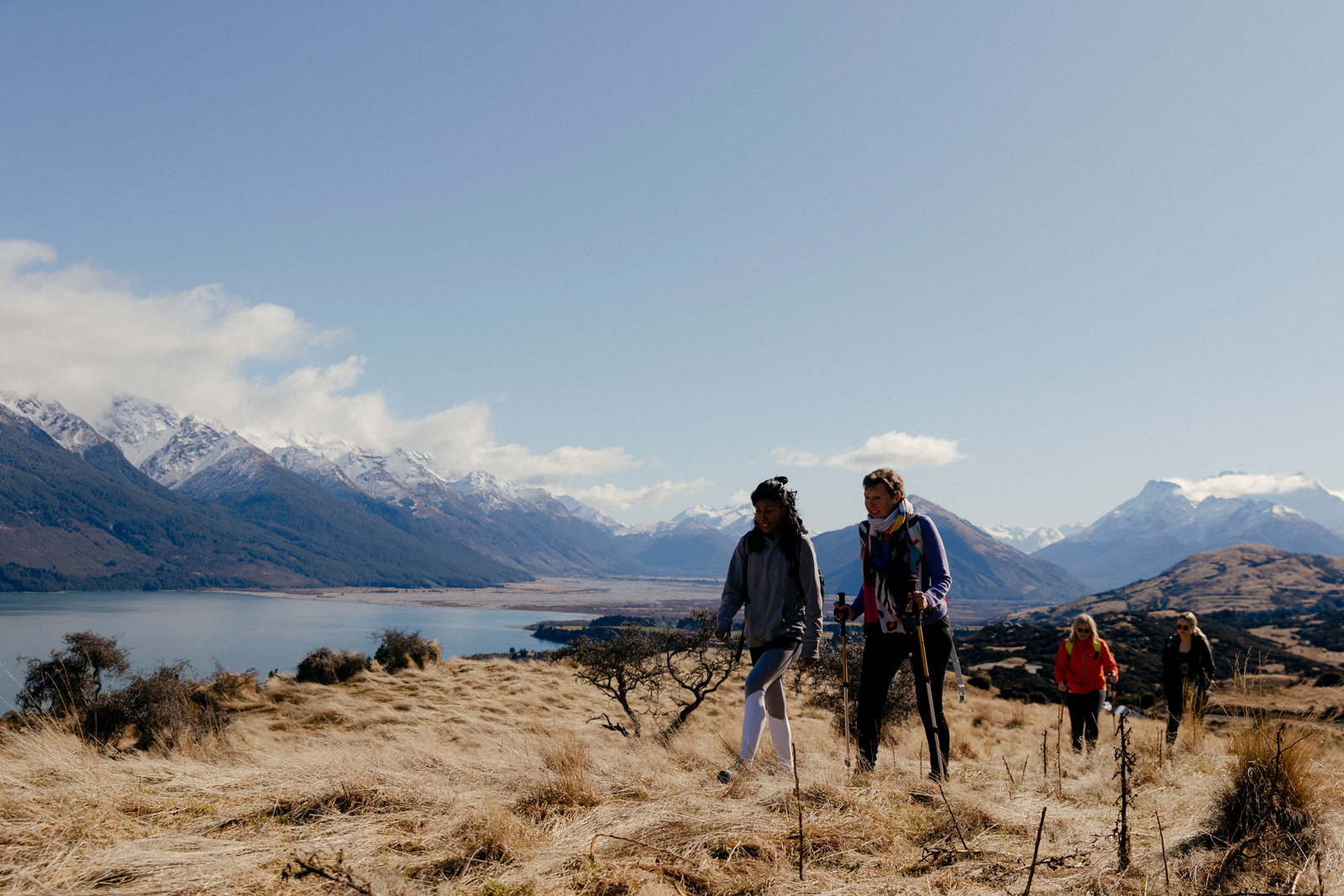 Two hikers trekking through a scenic mountain landscape with snow-capped peaks, a lake, and golden grasslands in the background.