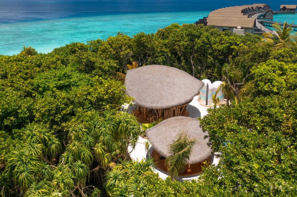 An aerial view of a lush tropical resort with thatched-roof villas nestled among green vegetation, overlooking turquoise blue ocean waters. The resort features unique circular bungalows with natural materials blending into the landscape.