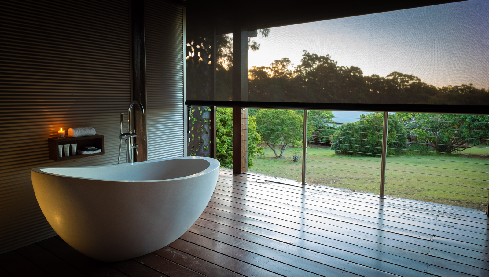 A modern, minimalist bathroom featuring a freestanding white bathtub on a wooden deck, overlooking a lush green garden at sunrise or sunset. Candles and towels add a spa-like ambiance to the tranquil space.