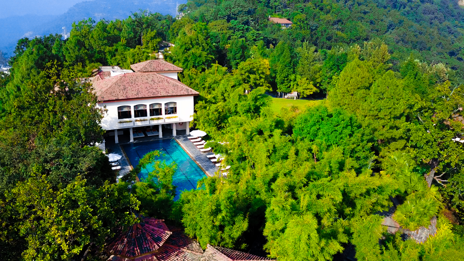 An elegant white villa with a large blue swimming pool nestled among lush green tropical trees and vegetation. The property features a terracotta-tiled roof and appears to be situated on a mountainous, forested landscape.