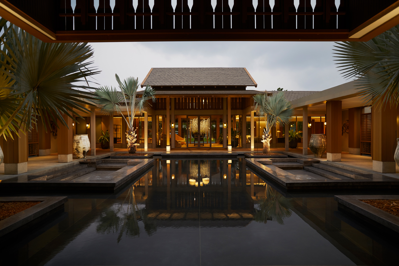 RAKxa-CHA-1 An elegant tropical resort architectural design featuring a central pavilion with symmetrical wings, surrounded by palm trees and a serene reflection pool illuminated during twilight hours.