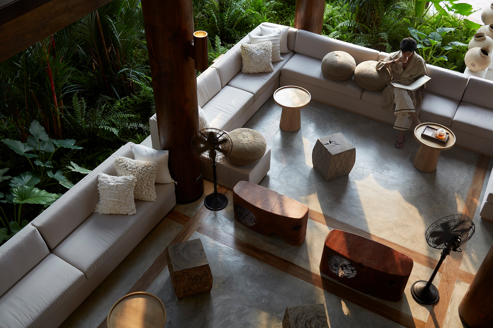 RAKxa-Jai-Lobby- A contemporary interior living space with minimalist white sofas, wooden accents, and floor-to-ceiling windows overlooking a dense tropical forest environment. The space features organic design elements and natural materials, creating a serene indoor-outdoor connection.