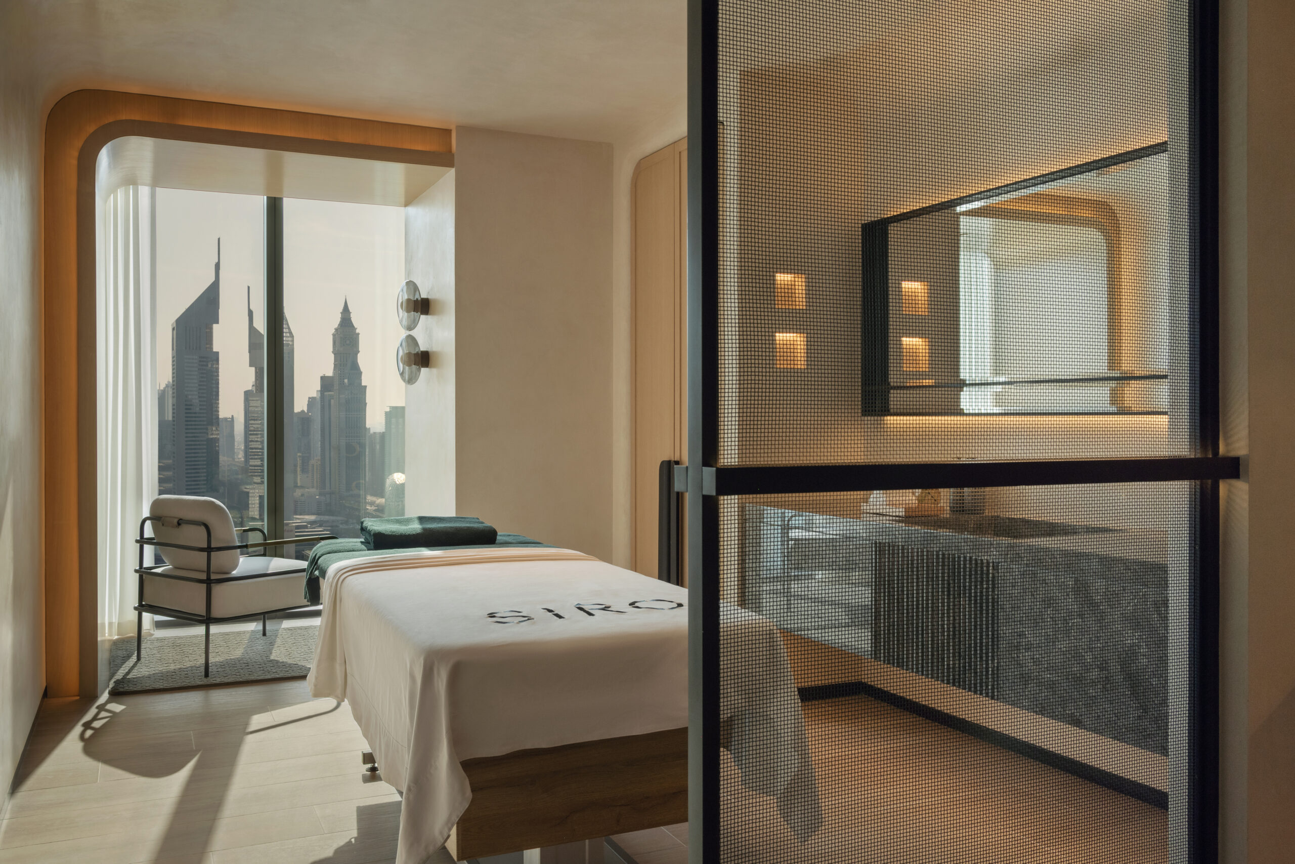 A modern spa treatment room with a white massage table and panoramic windows overlooking the distinctive skyscrapers of Dubai. The room features sleek design elements and a mesh screen partition.