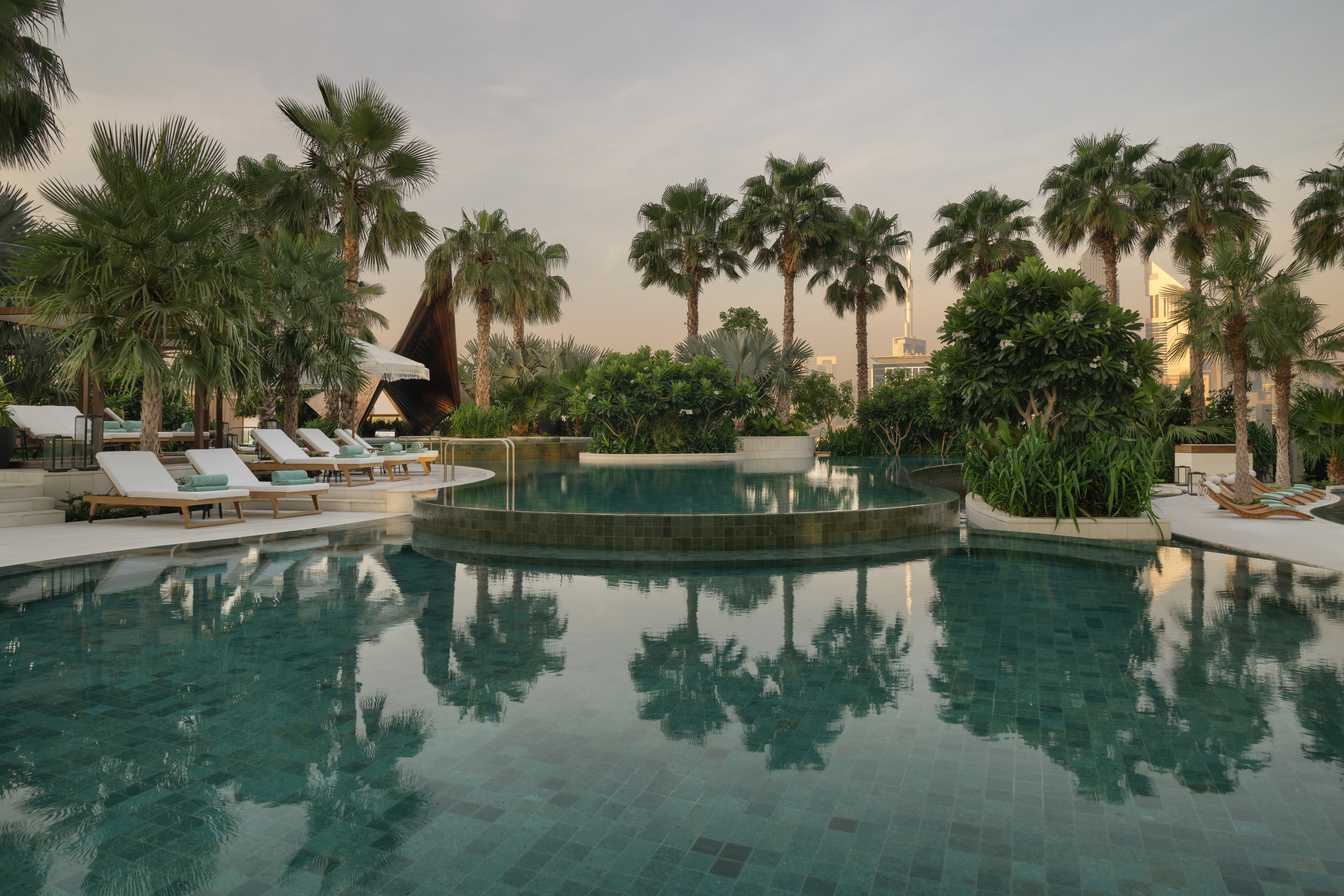 A serene resort swimming pool surrounded by lush palm trees and loungers, with a calm reflective surface creating a tranquil tropical atmosphere during golden hour.
