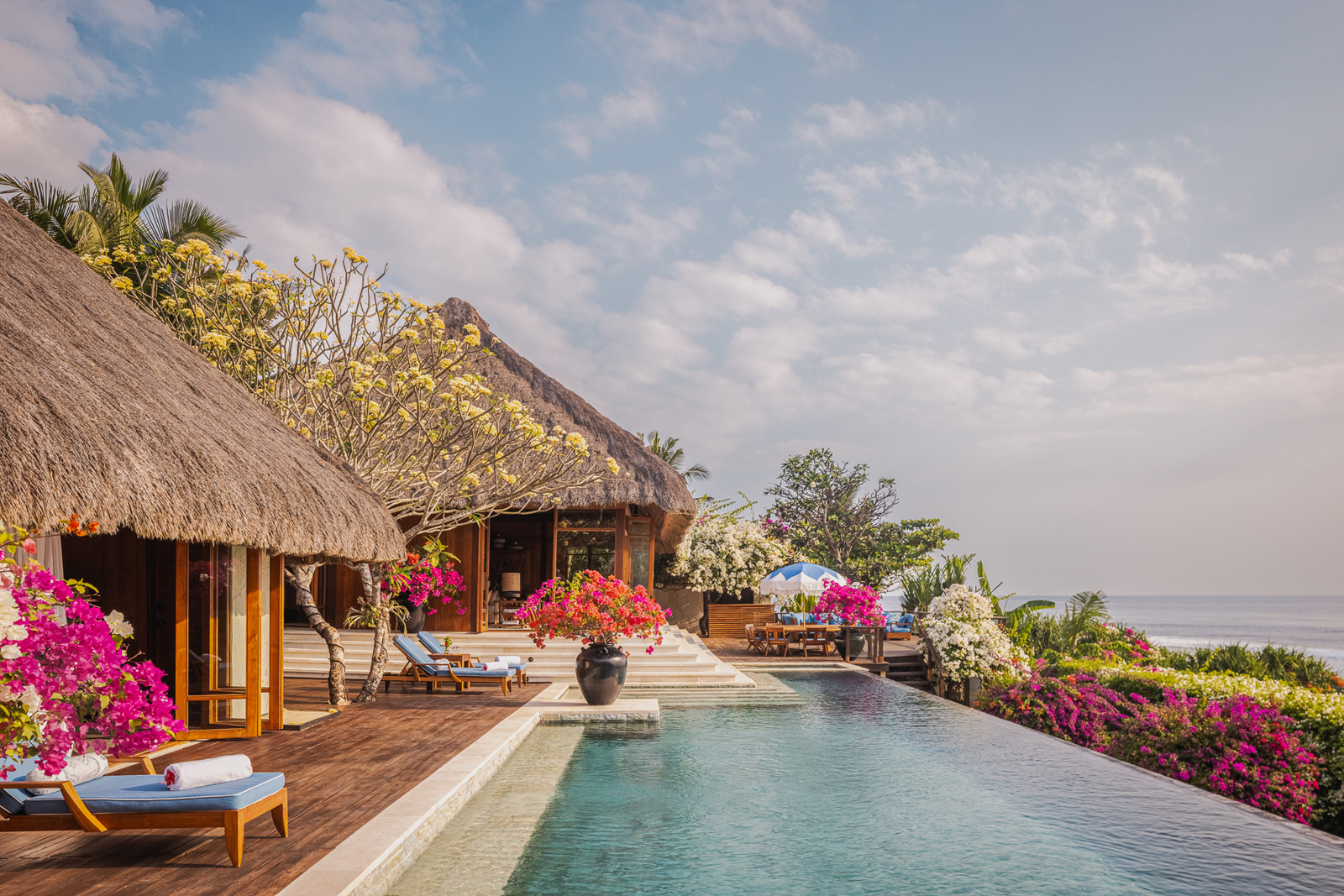 A stunning tropical resort villa with a thatched roof, featuring an expansive infinity pool overlooking the ocean, surrounded by vibrant bougainvillea and lush greenery.