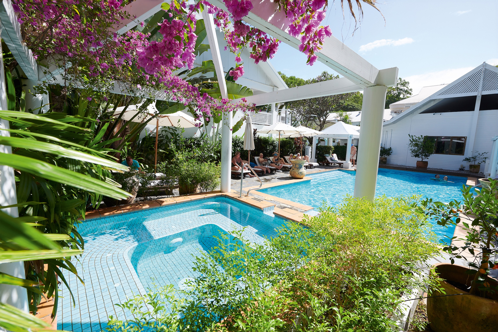 A luxurious swimming pool area with white pergola, lush greenery, and vibrant pink bougainvillea flowers. Guests relax on loungers under white umbrellas in a serene tropical setting.