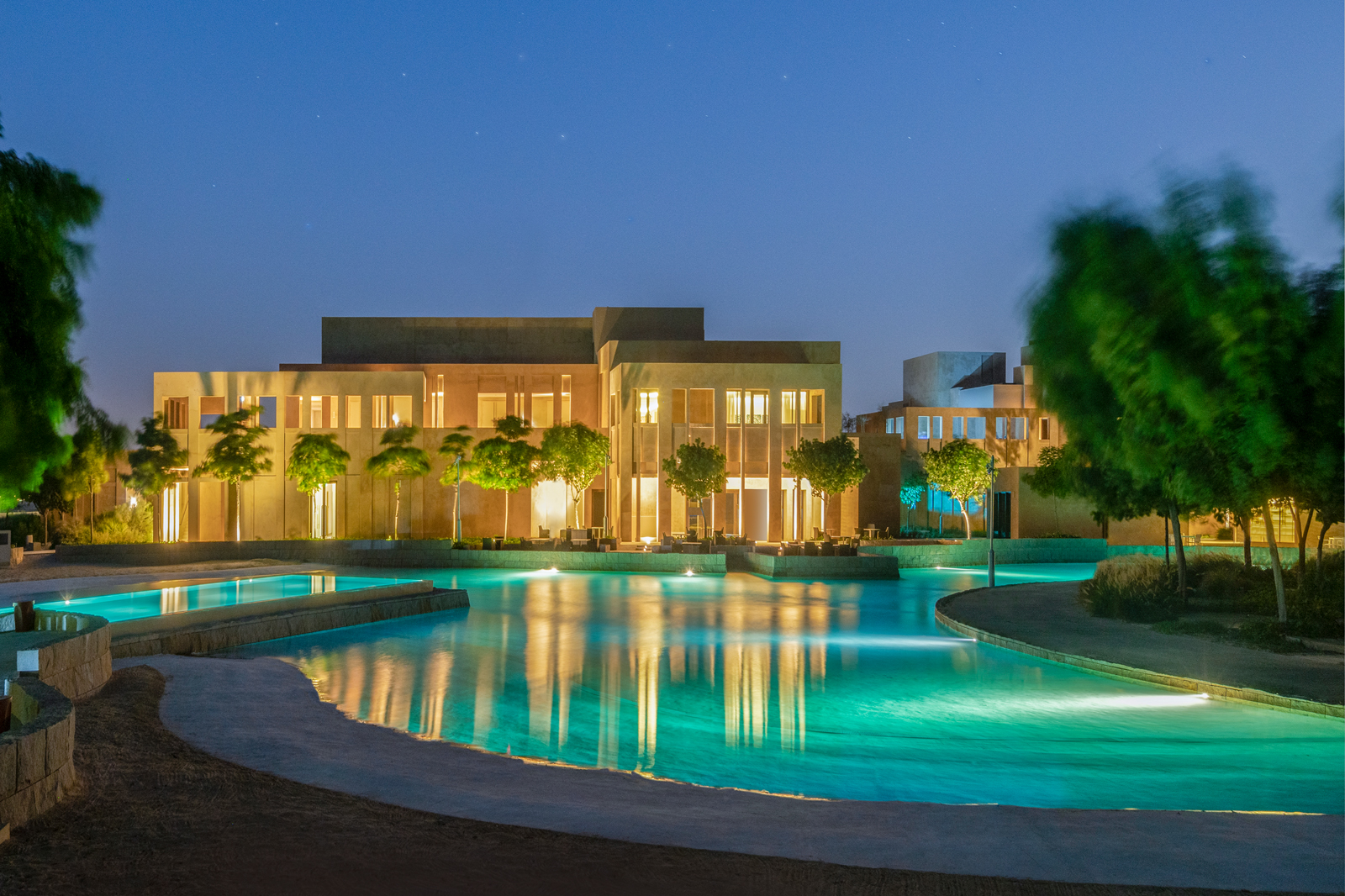 A stunning resort complex with illuminated buildings and a serene turquoise swimming pool surrounded by landscaped trees and gardens. The image captures the tranquil evening atmosphere with warm golden lighting and cool blue tones.