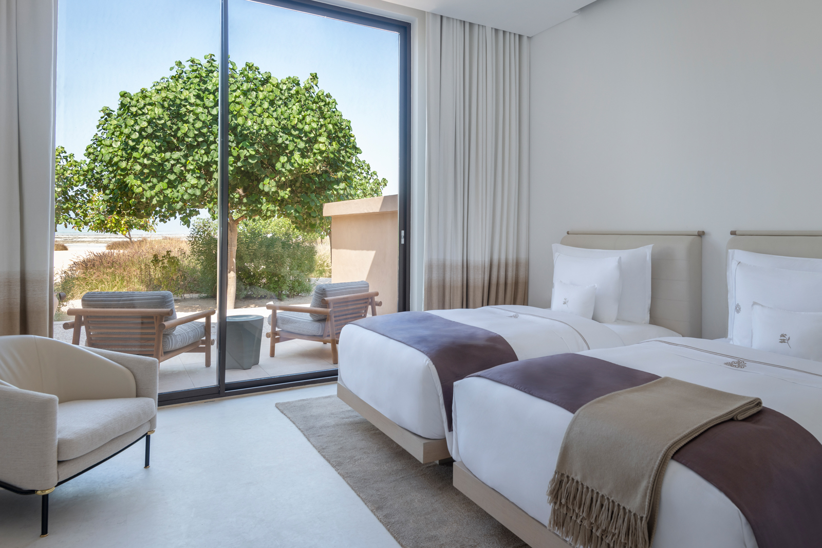 A modern, minimalist hotel bedroom featuring two twin beds with white linens and a large floor-to-ceiling window overlooking a lush green tree. The room has neutral tones and contemporary furnishings.