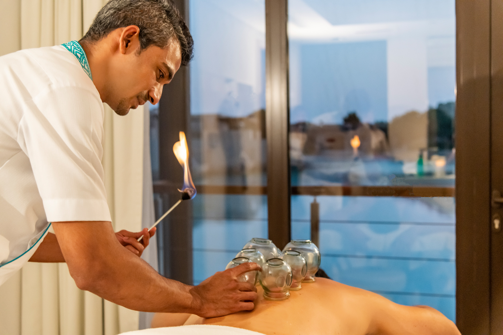 A practitioner performs cupping therapy, holding a flame to prepare glass cups for placement on a patient's back. The scene is set near a window with a view of the outside.