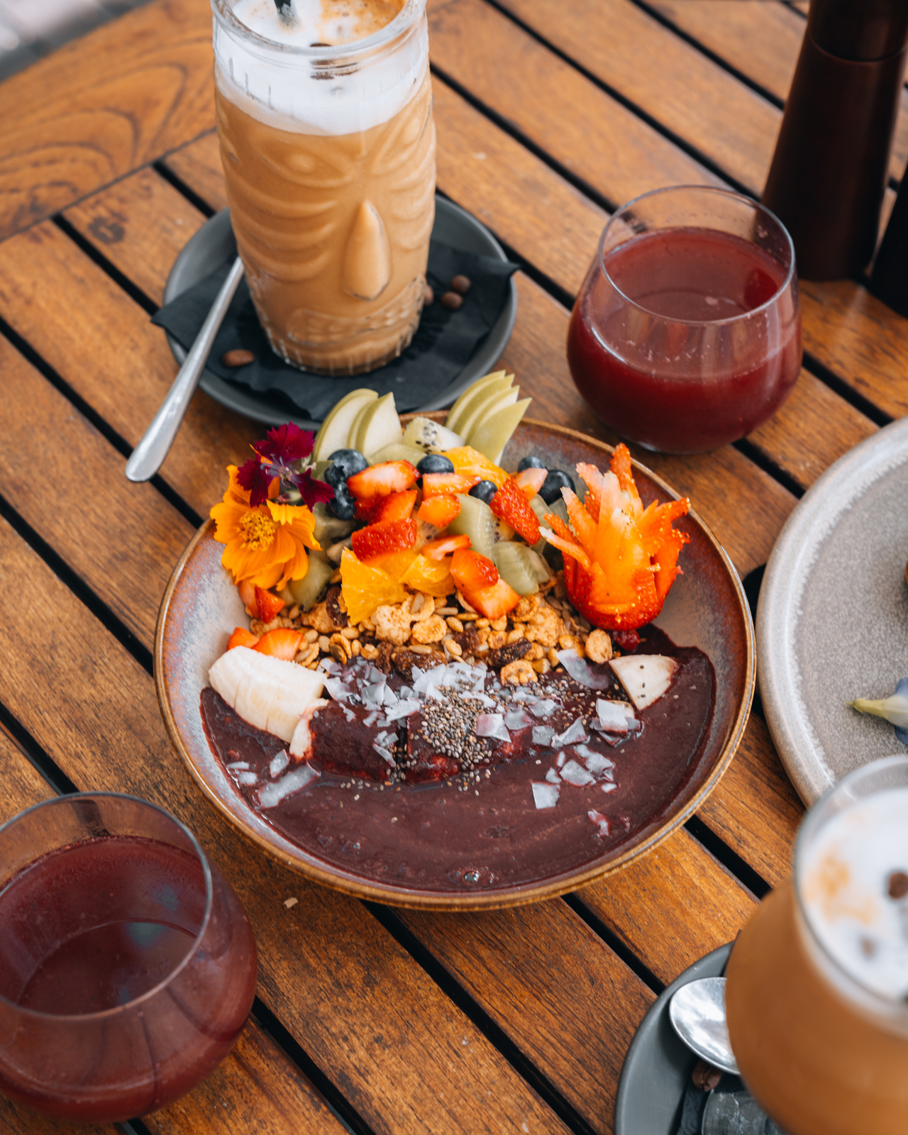 A beautifully composed açaí bowl with fresh fruits, granola, and chia seeds served alongside a latte and red juice on a wooden table.